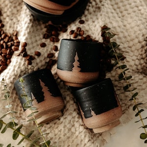 May include: Four black ceramic cups with a speckled finish and a brown base. Each cup features a tree silhouette. Coffee beans and greenery are scattered around the cups on a cream-coloured surface.