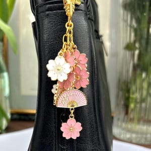 May include: A gold charm with pink and white cherry blossom flowers and a pink and gold fan charm. The charm is attached to a gold chain and is hanging from a black purse.