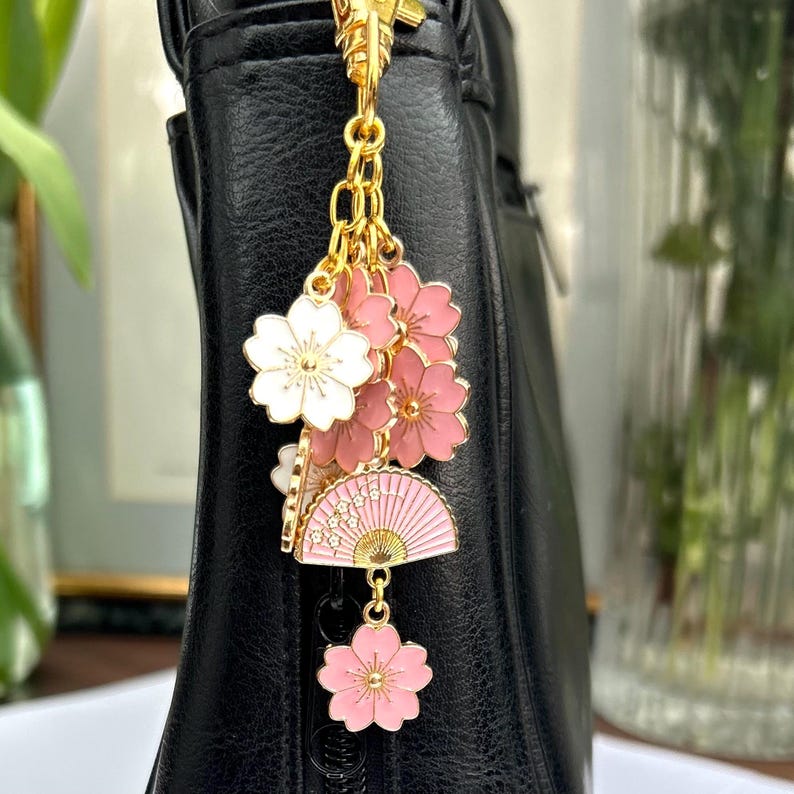 May include: A gold charm with pink and white cherry blossom flowers and a pink and gold fan charm. The charm is attached to a gold chain and is hanging from a black purse.