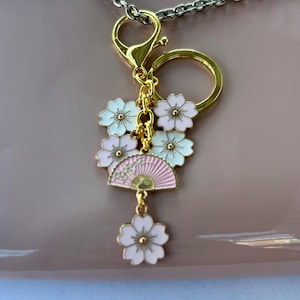 May include: A gold-colored keychain with a lobster clasp and a split ring. The charm features a pink fan and white and pink cherry blossom flowers. The keychain is attached to a silver-colored chain.
