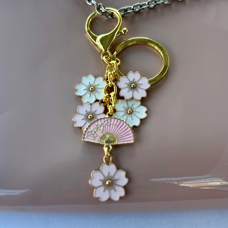 May include: A gold-colored keychain with a lobster clasp and a split ring. The charm features a pink fan and white and pink cherry blossom flowers. The keychain is attached to a silver-colored chain.
