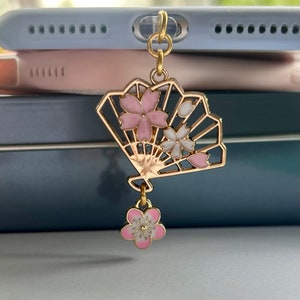 May include: A gold charm with a pink and white cherry blossom fan design. The charm has a small pink cherry blossom dangling from the bottom.
