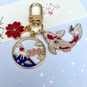 May include: Gold-tone keychain with Japanese-themed charms. Includes a red cherry blossom, a koi fish with red accents, and a circular charm with Mount Fuji, cherry blossoms, and clouds. The keychain has a lobster clasp.