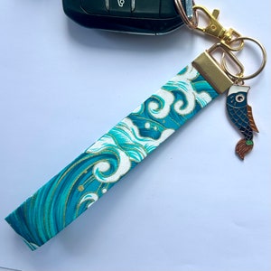 Peut inclure: Un porte-clés de poignet à motif de vagues turquoise et blanc avec une breloque poisson. Le tissu présente un motif de vagues avec des accents dorés. Le porte-clés est fixé à un fermoir et un anneau dorés.