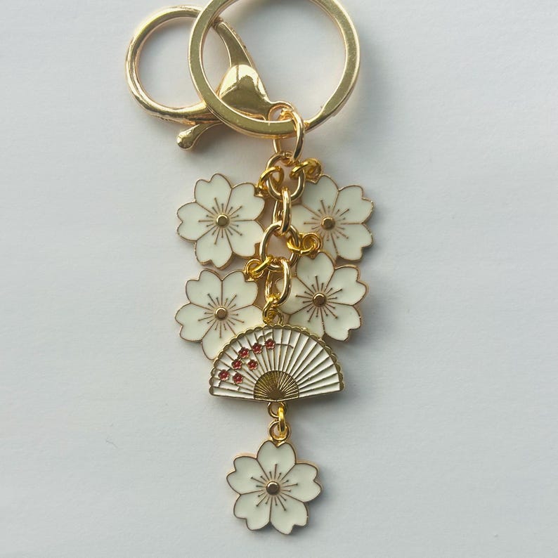 May include: Gold-tone keychain with white flower and fan charms. The charms are detailed with red accents. The keychain has a lobster clasp and a split ring for attaching keys.
