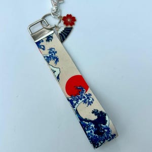 May include: A beige wrist strap keychain featuring a Japanese wave design in blue and a red sun. The keychain has a silver clasp and a charm with a red flower and a fan design.