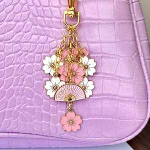 May include: A gold charm with pink and white cherry blossoms and a pink and gold fan. The charm is attached to a gold chain.