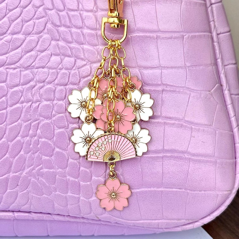 May include: A gold charm with pink and white cherry blossoms and a pink and gold fan. The charm is attached to a gold chain.