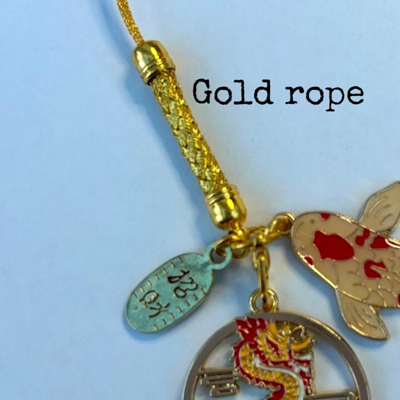 Op de afbeelding: Gouden touwtjes sleutelhanger met een rode en witte koi-vis charme, een ronde charme met een gouden draak en een kleine ovale charme met Chinese karakters.