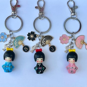 May include: Three keyrings with silver clasps. Each keyring features a Japanese Kokeshi doll with a black bob, a bow, and a kimono. Charms include flowers, fans, fish, and initial charms. The dolls wear blue, black, and pink kimonos.
