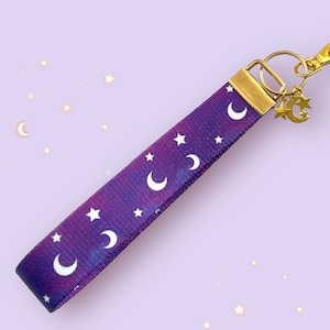 May include: Purple and white key chain with a galaxy print featuring white moons and stars. The key chain has a gold metal ring and a gold metal charm with a moon and star.
