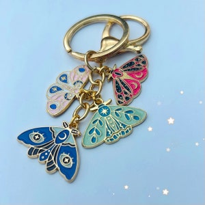 May include: A gold key chain with four enamel charms shaped like moths. The moths are decorated with celestial patterns and colours including pink, blue, green, and black.
