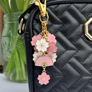 May include: A gold charm with pink and white cherry blossoms and a pink and gold fan. The charm is attached to a black purse with a gold chain.