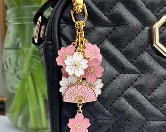 Könnte beinhalten: Ein goldener Anhänger mit rosa und weißen Kirschblüten und einem rosa und goldenen Fächer. Der Anhänger ist an einer schwarzen Handtasche mit einer goldenen Kette befestigt.