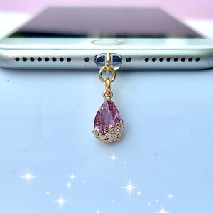 May include: A pink crystal teardrop charm with gold accents hangs from a gold chain. The charm is attached to a phone case.