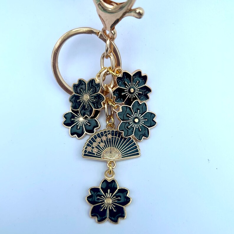 May include: A gold-colored keychain with a cluster of black flower charms and a fan charm. The charms are detailed with gold accents. The keychain has a lobster clasp and a ring for keys.