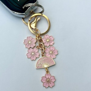 May include: A gold-colored keychain with a cluster of pink cherry blossom charms and a pink fan charm. The fan charm has Japanese characters. The keychain is attached to a car key.