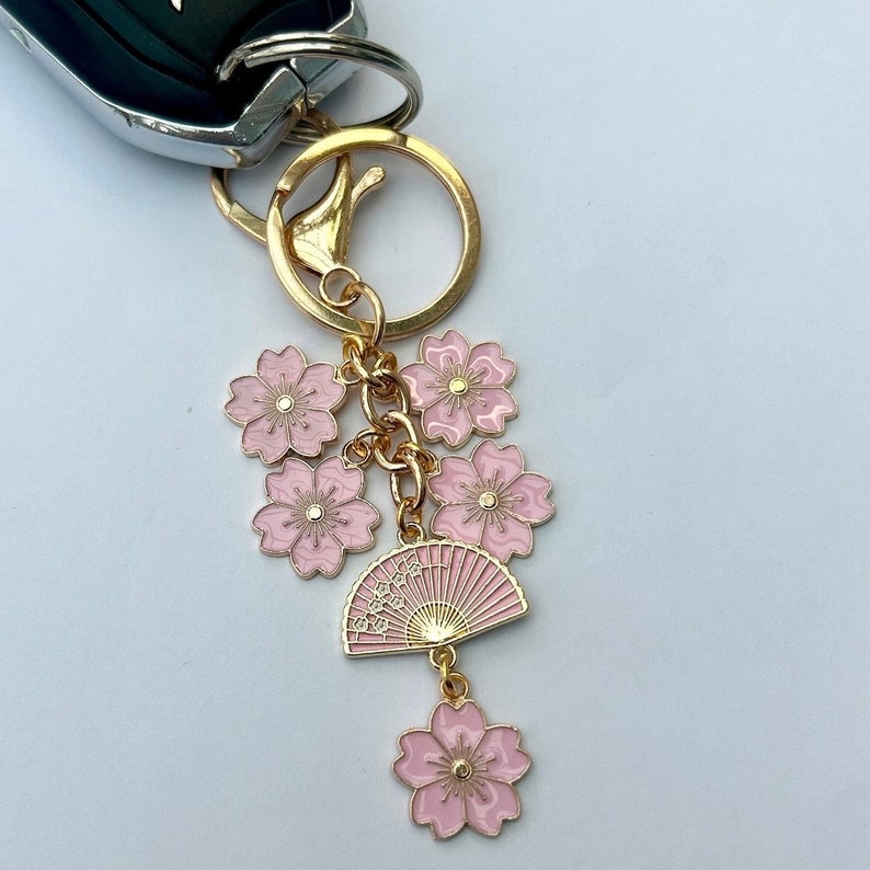 May include: A gold-colored keychain with a cluster of pink cherry blossom charms and a pink fan charm. The fan charm has Japanese characters. The keychain is attached to a car key.