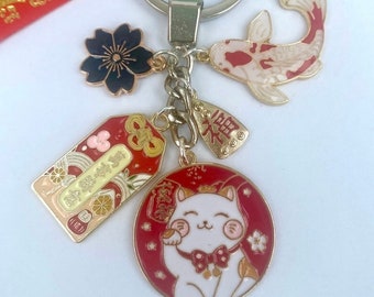 May include: A gold keychain with a red and white lucky cat charm, a red and gold tag with Chinese characters, a black and gold flower charm, and a red and white koi fish charm.