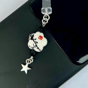 May include: A white ceramic charm with black and red details, a black bead, and a silver star charm attached to a silver chain. The charm is hanging from a clear plastic phone charm holder.