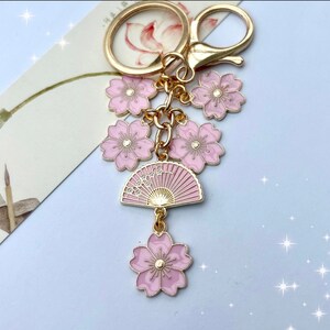 May include: A gold-colored keychain with pink cherry blossom flower charms and a pink fan charm. The keychain has a lobster clasp and a key ring. The fan charm has a gold-colored design. The flowers have gold-colored accents.