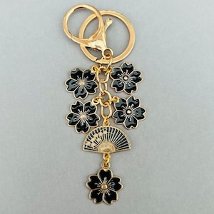 May include: A gold-colored keychain featuring black floral and fan charms. The charms are connected by gold-colored chains and a clasp. The keychain has a ring for keys.