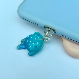 May include: A blue whale shark phone charm with white spots, attached to a light blue phone case via a silver chain. The phone case is light blue. The charm is a decorative accessory.