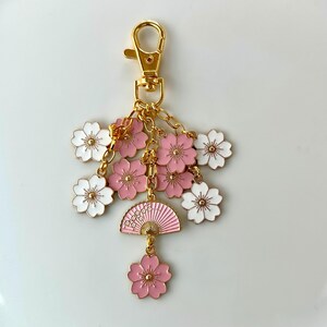 May include: Gold keychain with a pink and white fan charm and multiple pink and white flower charms. The fan charm has a gold design and the flower charms have gold accents.