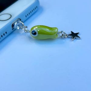May include: A lime green phone charm with a fish-shaped bead and a silver star charm. The fish bead has a large, blue eye. The charm is connected to a phone via the headphone jack.