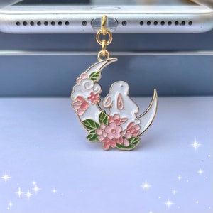 May include: A gold-tone charm featuring a white crescent moon with a white rabbit sitting on it. The moon is decorated with pink and green enamel flowers.