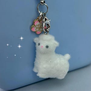 May include: A white, fluffy sheep charm with a pink nose and black eyes, attached to a silver clasp and a pink and gold flower charm. The background is a light blue.