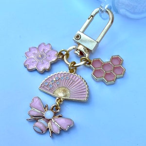 May include: A gold keychain with pink and white enamel charms. The charms include a bee, a fan, a honeycomb, and a flower.