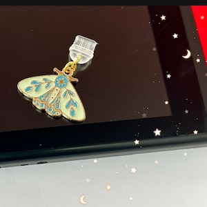 May include: A gold-colored charm with a light blue and green moth design. The charm has a clear plastic loop for attaching to a bag or keychain.