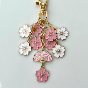 May include: A gold keychain with a pink and white cherry blossom design. The keychain features a small fan charm and multiple flower charms.