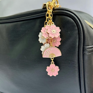May include: A gold charm with pink and white flowers and a pink and gold fan charm. The charm is attached to a gold chain and is hanging from a black leather bag.