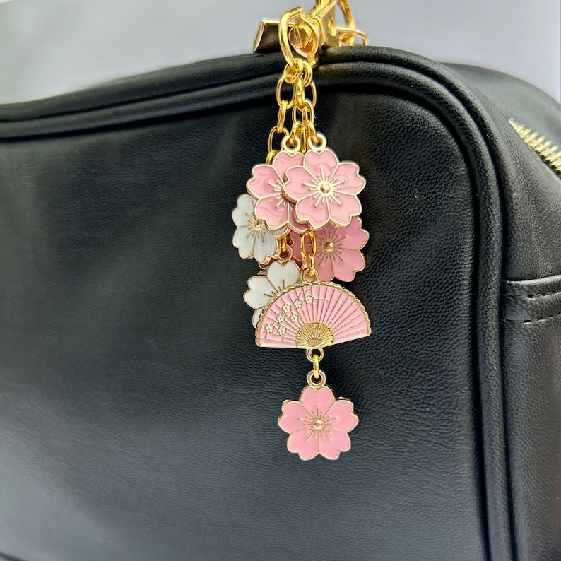 May include: A gold charm with pink and white flowers and a pink and gold fan charm. The charm is attached to a gold chain and is hanging from a black leather bag.
