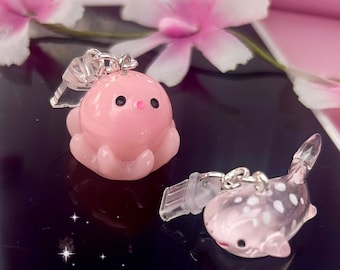 Puede incluir: Dos colgantes rosas translúcidos: un pulpo y un pez. El pulpo tiene ojos negros y un pico pequeño. El pez tiene manchas blancas y una boca roja. Ambos colgantes tienen accesorios plateados.