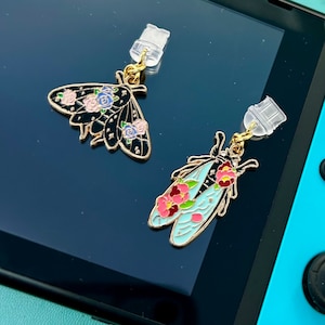 May include: Two charm pendants featuring detailed butterfly designs with floral accents. The butterfly on the left is black with blue and pink flowers, while the butterfly on the right is blue with pink flowers. Both pendants have gold accents and clear plastic attachments.