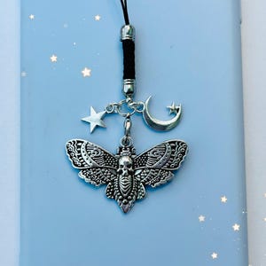 May include: A silver charm featuring a detailed moth design, a skull, and a crescent moon with a star. The charm is attached to a black cord and hangs from a phone case.
