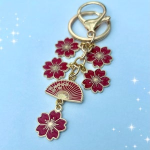 May include: A gold-toned keychain featuring a cluster of red cherry blossom charms and a red fan charm. The keychain has a lobster clasp and a key ring. The blossoms have gold accents.