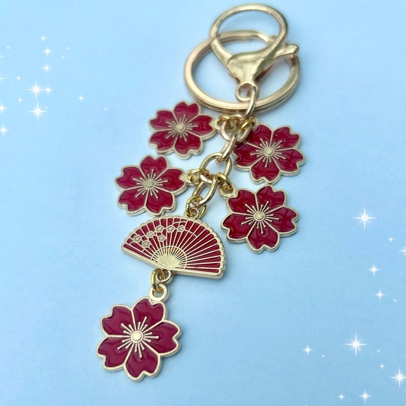 May include: A gold-toned keychain featuring a cluster of red cherry blossom charms and a red fan charm. The keychain has a lobster clasp and a key ring. The blossoms have gold accents.