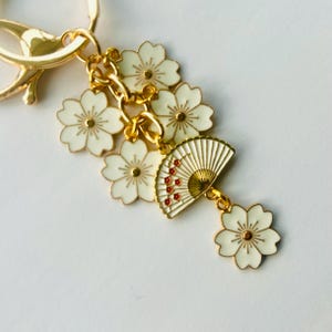 May include: Gold-tone keychain with white flower and fan charms. The charms feature white enamel with gold accents and red details on the fan. The keychain has a lobster clasp for easy attachment.
