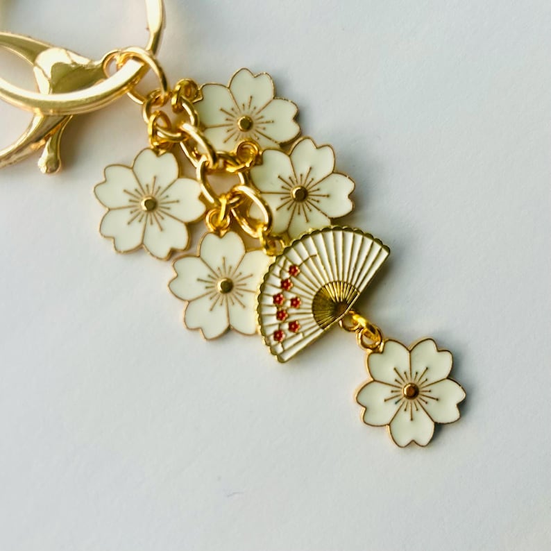 May include: Gold-tone keychain with white flower and fan charms. The charms feature white enamel with gold accents and red details on the fan. The keychain has a lobster clasp for easy attachment.