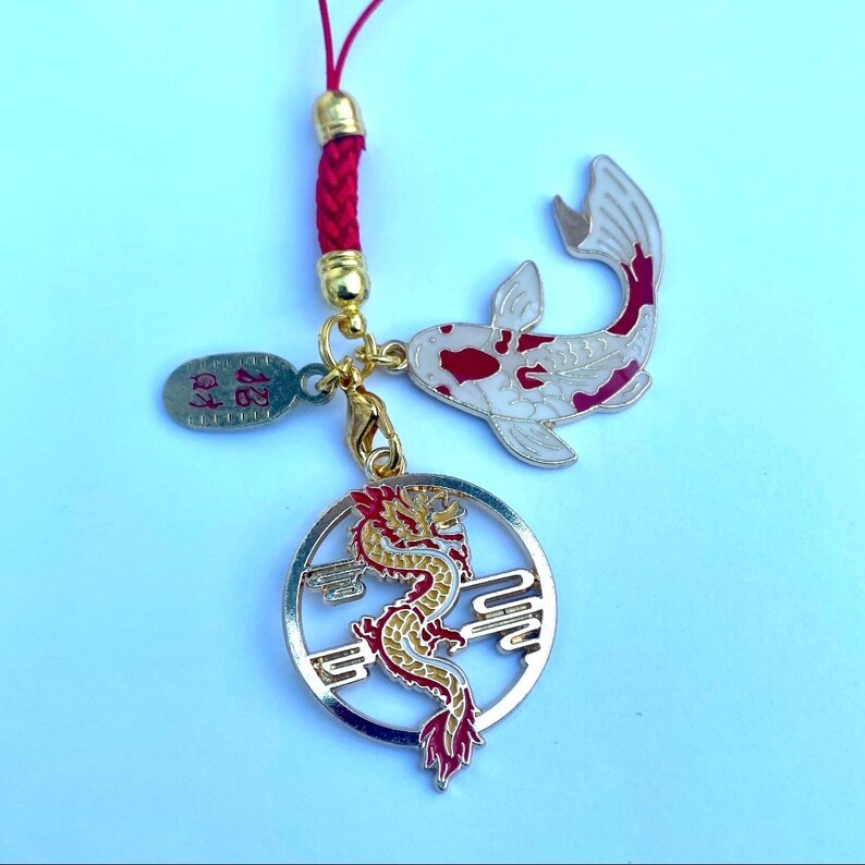Op de afbeelding: Een rode en gouden hanger met een rode draak en een witte koi-vis. De hanger is bevestigd aan een rood touwtje.