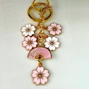 May include: A gold-colored keychain with a cluster of pink and white flower charms and a pink fan charm. The charms are connected by gold-colored chains and a clasp. The flowers have gold-colored centers.