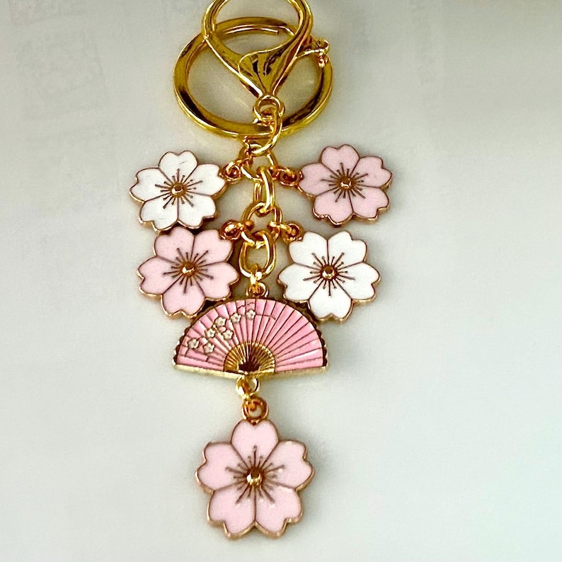 May include: A gold-colored keychain with a cluster of pink and white flower charms and a pink fan charm. The charms are connected by gold-colored chains and a clasp. The flowers have gold-colored centers.