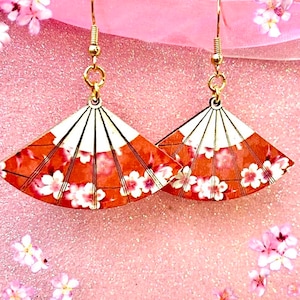 May include: A pair of gold earrings featuring a fan-shaped design. The fans are crafted from wood and decorated with a cherry blossom pattern in shades of pink and orange.