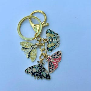May include: A gold-colored keychain with four enamel butterfly charms in various colors. The butterflies are yellow, black, pink, and teal, with intricate designs. The keychain has a lobster clasp and a split ring.