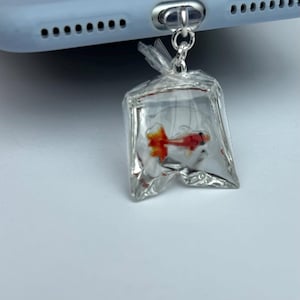 May include: A silver phone charm depicting a goldfish in a clear plastic bag. The goldfish has orange and red fins and tail. The charm is attached to a phone case.