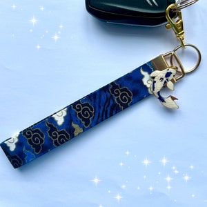May include: Blue and gold key chain with a white koi fish charm. The key chain has a blue fabric strap with a pattern of white clouds and gold accents.